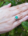 Hand wearing an emerald ring with a green leafy background.