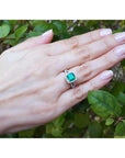 Hand wearing an emerald ring with a green leafy background.