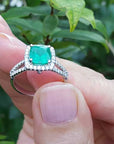 Held between fingers a bridal's emerald engagement ring with a green leafy background.