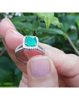 Held between fingers a bridal's emerald engagement ring with a green leafy background.