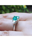 2.18 ct cushion cut emerald ring with trillion side diamonds in 14K white gold on a finger.