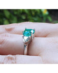2.18 ct cushion cut emerald ring with trillion side diamonds in 14K white gold on a finger.