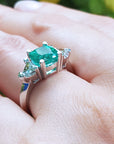 May's birthstone emerald ring cushion cut trillion side diamonds hand against a blurred natural background.