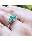 May's birthstone emerald ring cushion cut trillion side diamonds hand against a blurred natural background.