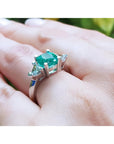 2.18 ct cushion cut emerald ring with trillion side diamonds on a hand, 14K white gold.
