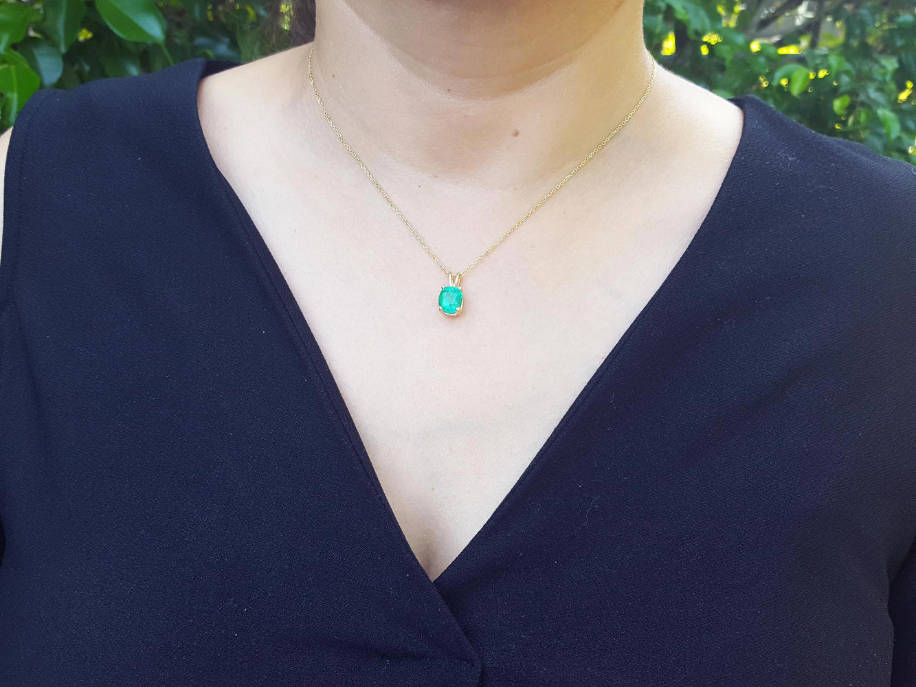 A woman wearing a necklace with a cushion emerald pendant against a blurred green background.
