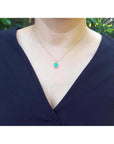 A woman wearing a necklace with a cushion emerald pendant against a blurred green background.