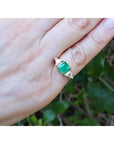 A 18k yellow gold emerald ring with trillion side diamonds on a hand against a natural background.
