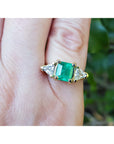 A 18k yellow gold genuine Muzo colombian emerald ring with trillion diamonds on a finger against a blurred natural background.