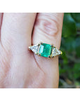 A 18k yellow gold genuine Muzo colombian emerald ring with trillion diamonds on a finger against a blurred natural background.