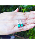A pair of emerald dangle earrings placed on a hand with a blurred green leafy background