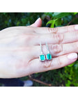 18K white gold dangling emerald-cut emerald earrings with diamond-studded lines on a hand.