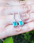 Pair of emerald-cut gemstone earrings held in a hand with a natural background