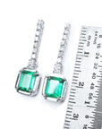 Pair of 18k white gold emerald dangle earrings next to a ruler for scale on a white background.
