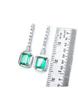 Pair of 18k white gold emerald dangle earrings next to a ruler for scale on a white background.