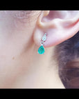Dangling teardrop emerald and diamond earrings in 14K white gold on an ear.