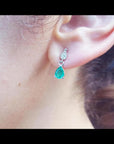Dangling teardrop emerald and diamond earrings in 14K white gold on an ear.