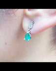 Dangling teardrop emerald earrings in 14K white gold with diamond-studded pear link on an ear.