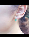 Dangling teardrop emerald earrings in 14K white gold with diamond accents on a woman's ear.