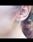 Dangling teardrop emerald earrings in 14K white gold with diamond accents on a woman's ear.