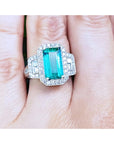 Anillo con esmeralda colombiana rectangular rodeada de diamantes en oro blanco exhibida entre dedos.