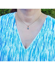 A woman wearing a blue and white patterned top with a necklace featuring a blue opal pendant.