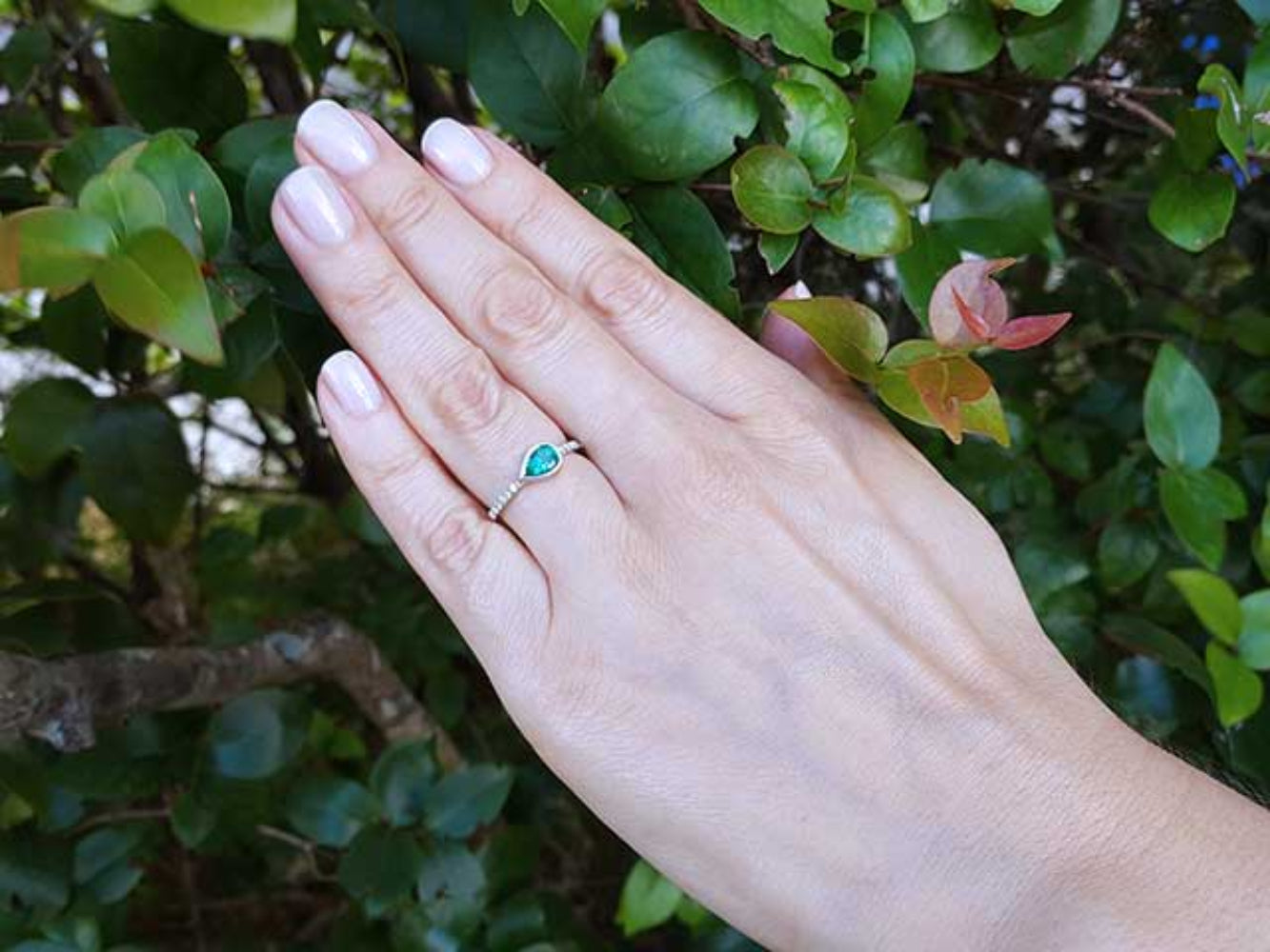 Hand wearing a ring with a teardrop natural emerald against a green leafy background.