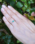 Hand wearing a ring with a teardrop natural emerald against a green leafy background.