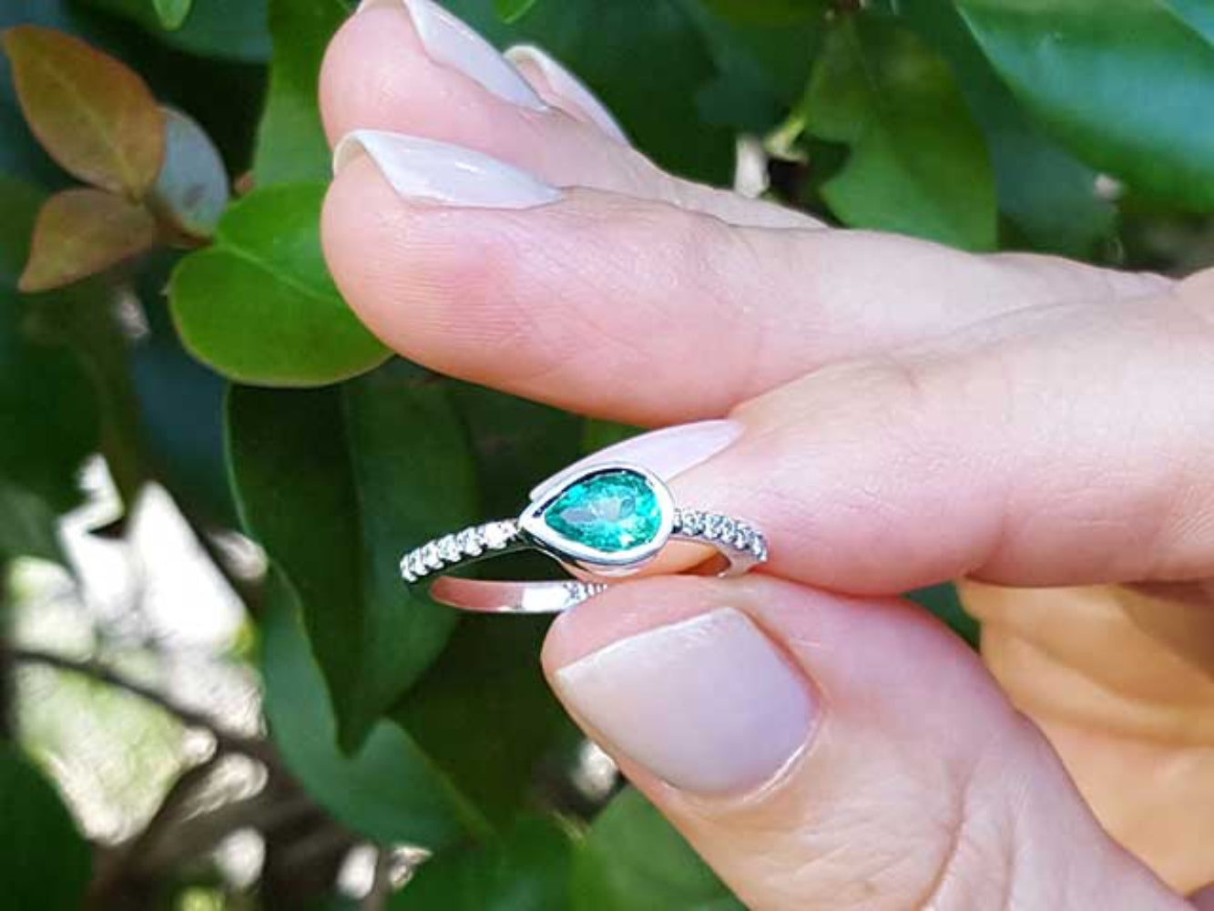 white gold ring with a teardrop natural emerald held between fingers against a blurred green leafy background.