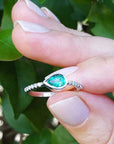 white gold ring with a teardrop natural emerald held between fingers against a blurred green leafy background.