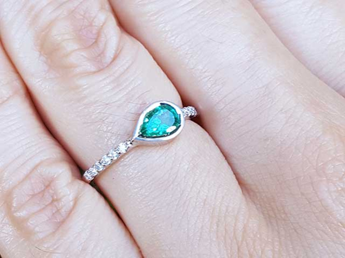 A teardrop emerald bezel set ring with accent diamonds in white gold on a finger.