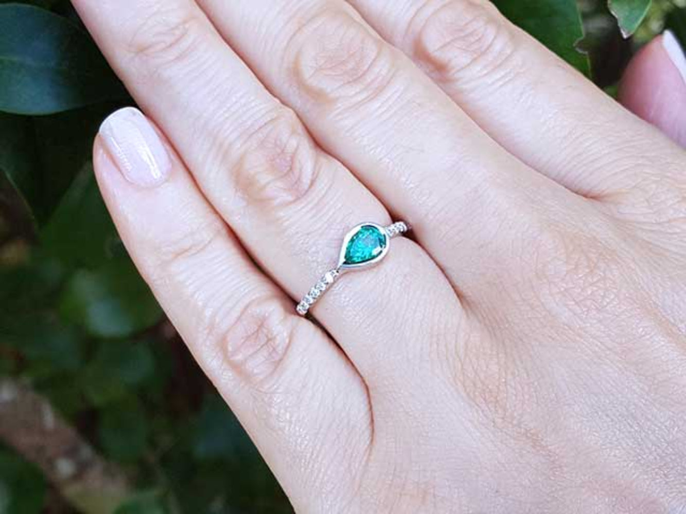 Hand wearing a ring with a bezel set natural colombian emerald against a blurred green background.