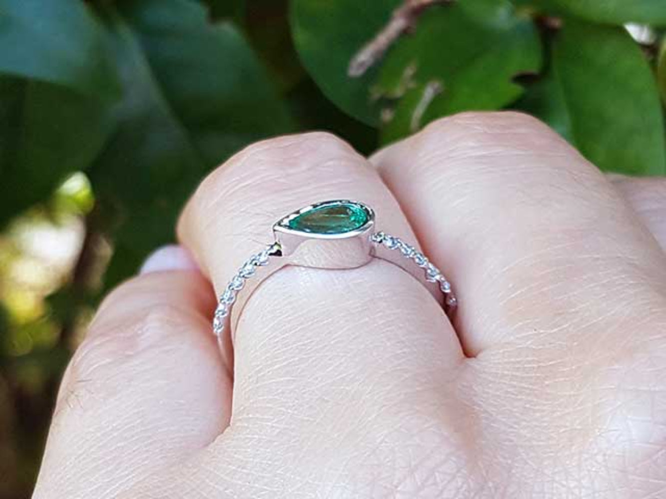 A white gold ring with a natural colombian emerald on a finger against a leafy background.
