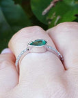 A white gold ring with a natural colombian emerald on a finger against a leafy background.