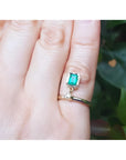14K yellow gold dangle ring with a bezel-set emerald and small diamond on a finger.
