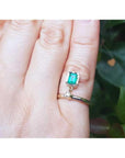 14K yellow gold dangle ring with a bezel-set emerald and small diamond on a finger.