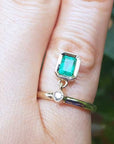 A yellow gold emerald dangle ring on a finger against a blurred natural background.