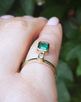 An emerald dangle ring representing May's birthstone jewelry on a hand against a blurred green leafy background.