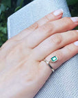 Hand wearing an emerald dangle ring against a blurred natural background.