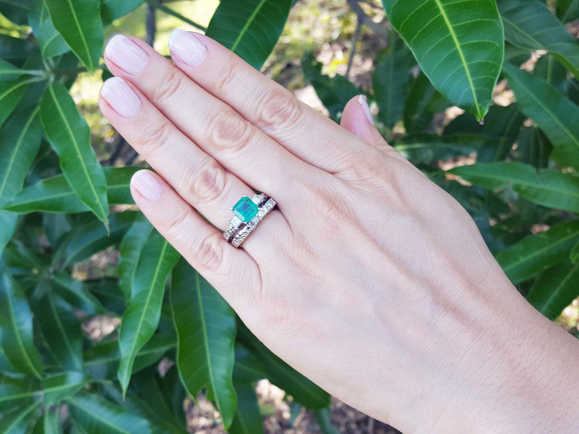 Hand wearing an emerald ring with a match band against a green leafy background.