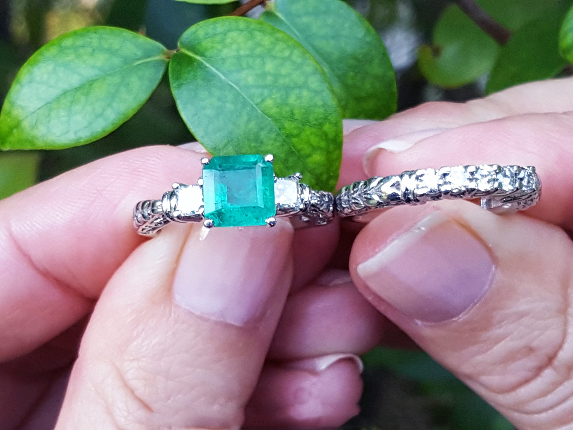 Between fingers holding an emerald ring and its matching diamond wedding band on a leafy background.