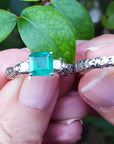 Between fingers holding an emerald ring and its matching diamond wedding band on a leafy background.
