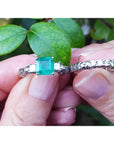 Between fingers holding an emerald ring and its matching diamond wedding band on a leafy background.