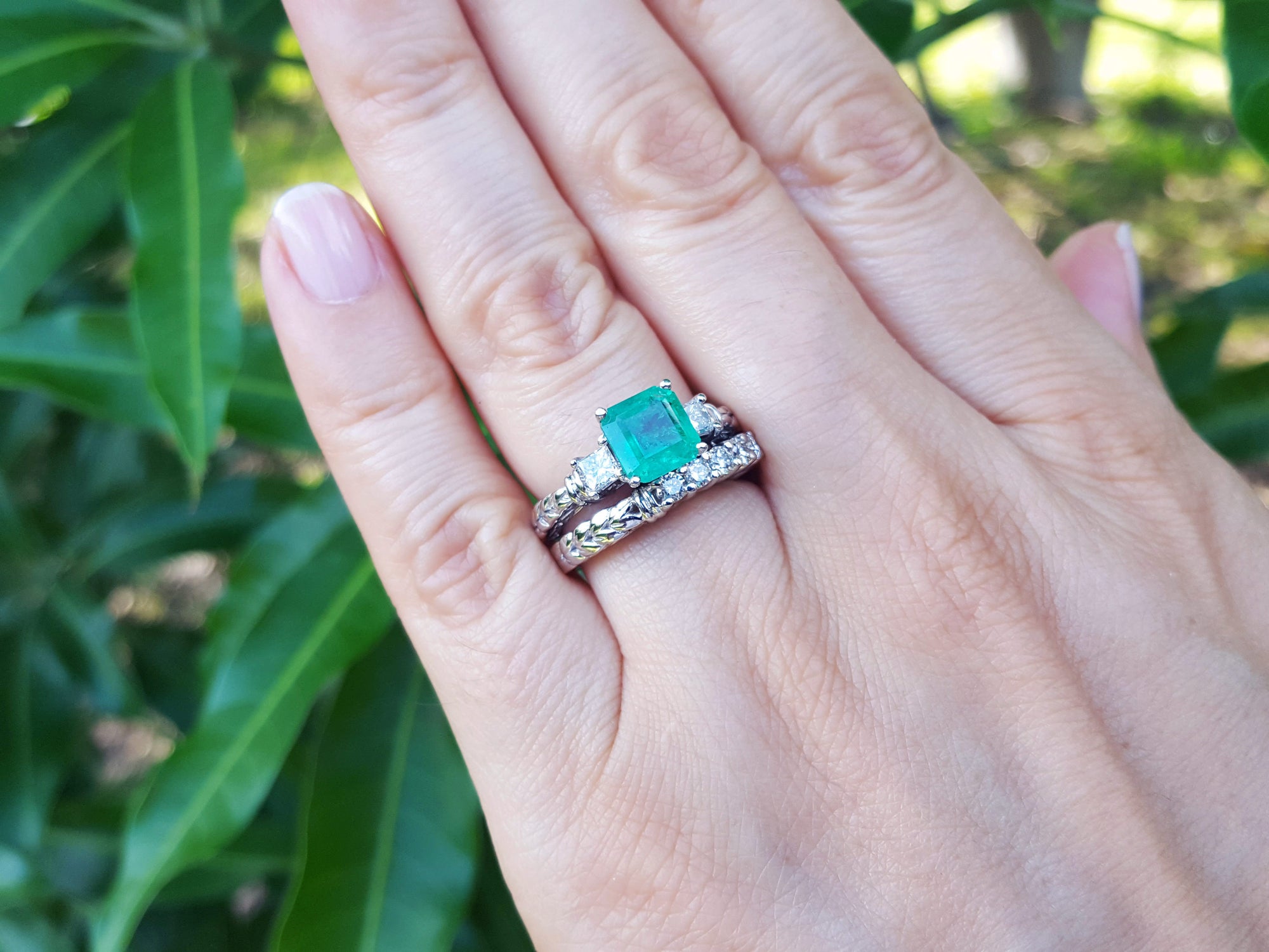 A close-up of a emerald and wedding band ring set on a green leafy background.