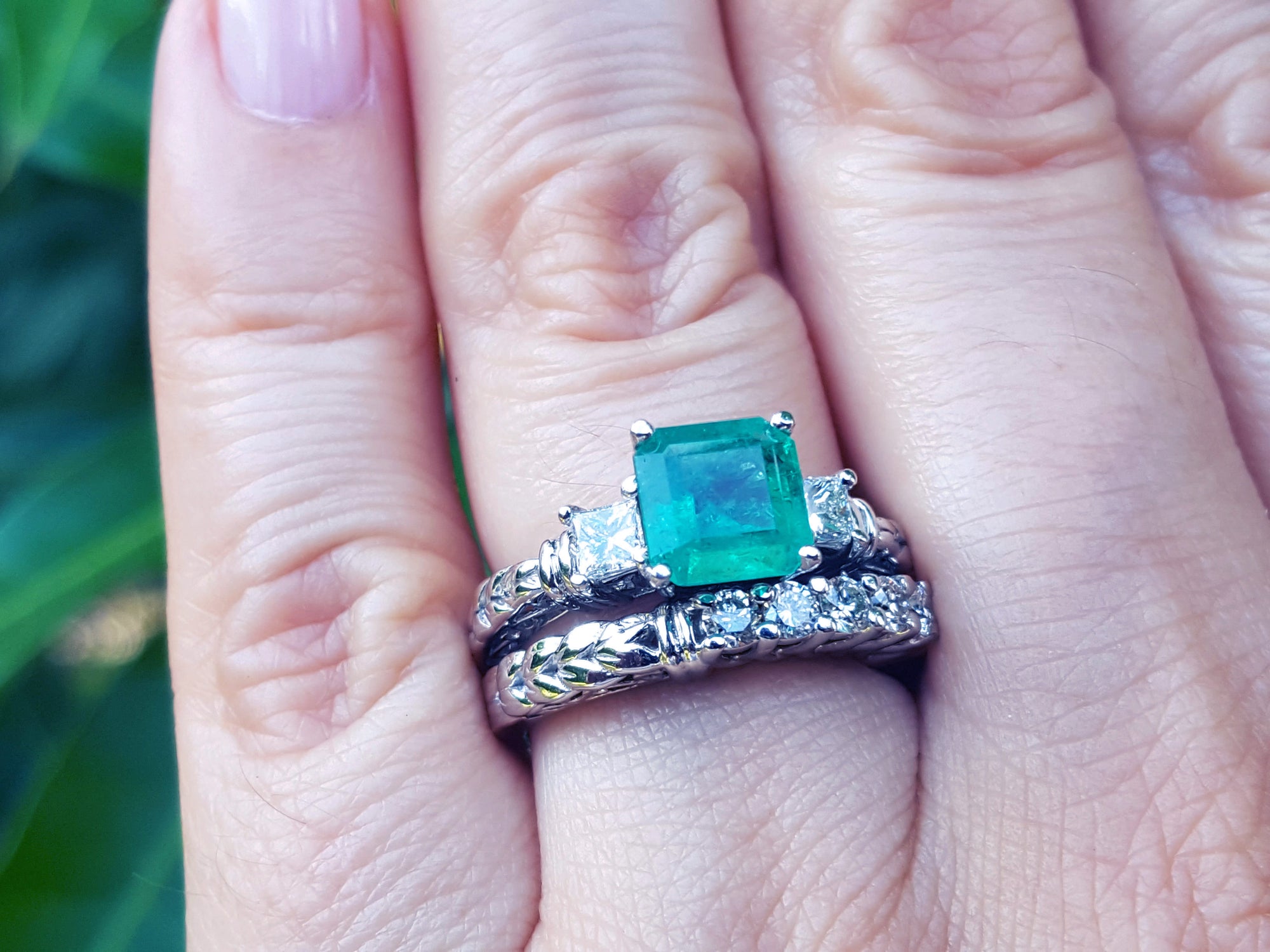 A hand wearing an emerald ring with matching wedding band on a green blurry background.