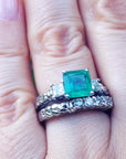 A hand wearing an emerald ring with matching wedding band on a green blurry background.