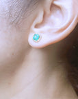 Close-up of a woman's ear wearing a green emerald stud earring with a blurred background.