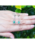Diamond-shaped earrings with marquise emerald gemstones on a hand against a green leafy background