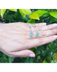 Hand displaying a pair of diamond-shaped emerald marquise earrings against a green leafy background.