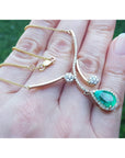 Hand holding a yellow gold necklace with a natural emerald gemstone pendant against a blurred green background